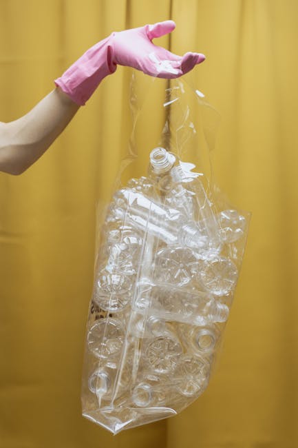 A middle-aged woman with short dark hair, wearing a black short-sleeved shirt with patterned text and dark pants, is bending over and placing a large white plastic bag filled with rubbish into a stain