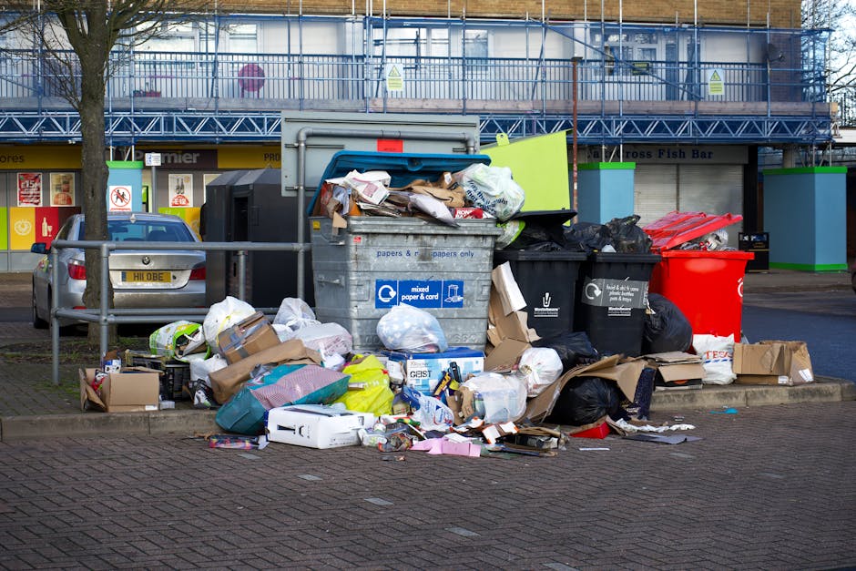 A man dressed in a red t-shirt, light blue denim shorts, and a black cap is standing on a sandy beach, holding a white rubbish bag in each hand as he participates in a litter cleanup activity. Beside 